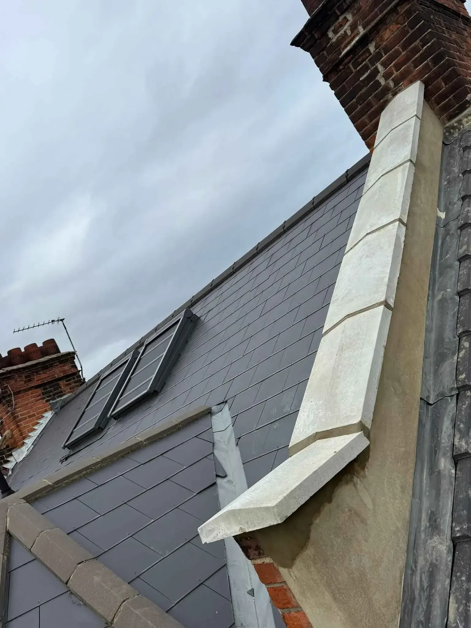 Rendered parapet wall with new pale capstones beside a brick chimney on a slate roof in Morden, Greater London